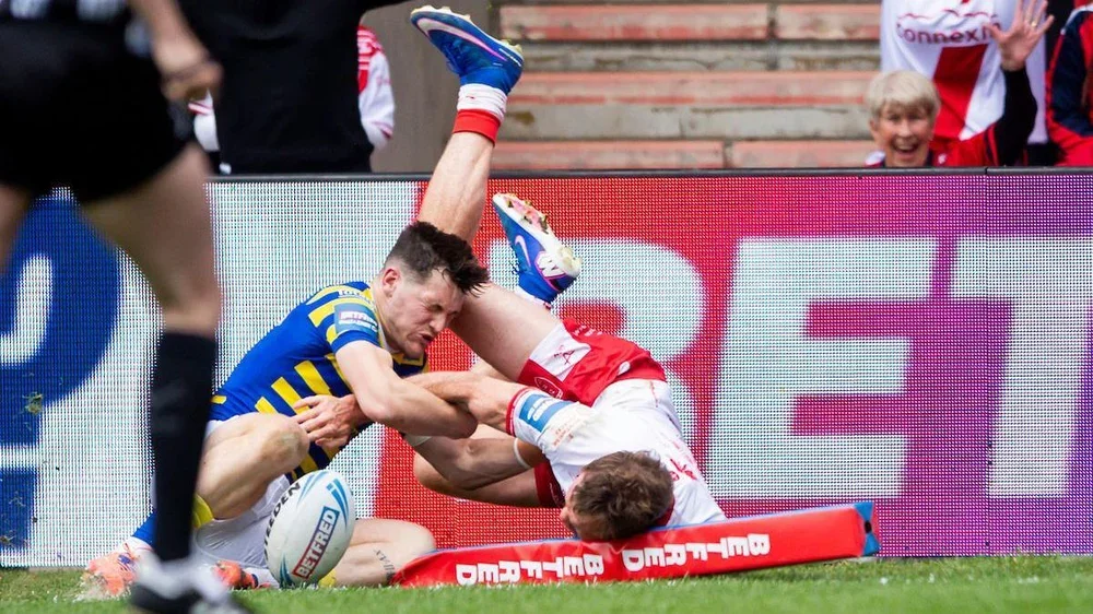 Hull KR player Joe Burgess, in white shirt and red shorts, flattens the corner post as he dives, legs in the air, after scoring a try as Warrington's Josh Thewlis, in blue and yellow hoops, tries to grab him